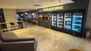 A modern lounge area with a sofa, a yellow chair, and a food and drink station with refrigerated displays. - Modern Flat in Gonzaga, just a block from the beach. (Santos)