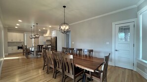A spacious dining area with a long wooden table and chairs, a kitchen in the background, and a chandelier.