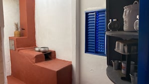 A kitchen with a red brick stove, a window with blue shutters, and shelves with kitchenware. - Fazenda Brasileiro (Passos)