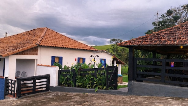 A white building with a red-tiled roof, a black gate, and a small garden with yellow flowers. - Fazenda Brasileiro (Passos)
