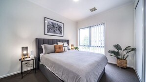 Une chambre à coucher moderne avec un grand lit, une table de chevet avec une lampe, un tableau encadré au mur et une fenêtre donnant sur l’extérieur.