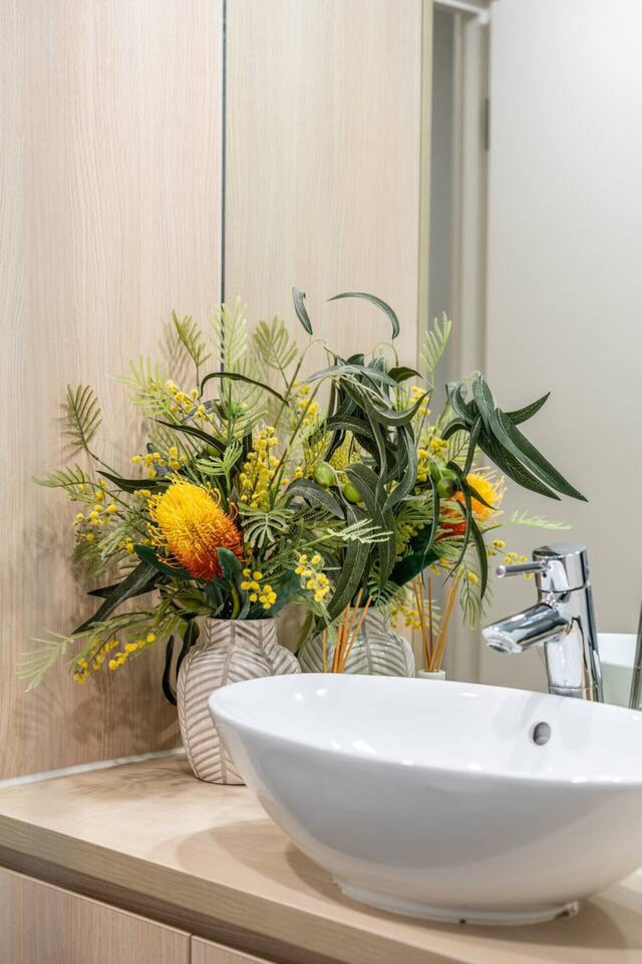 Un bagno con un lavandino bianco, uno specchio e un vaso con fiori gialli e verde decorativo.