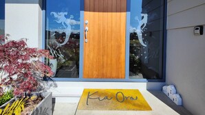 Una casa moderna con puerta principal de madera, ventana de vidrio con detalles decorativos y felpudo amarillo que dice "Kia Ora".
