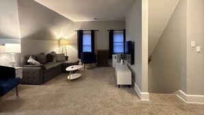 A living room with a sofa, a coffee table, a TV, and a staircase leading to another level. - NH3 - Historic Charm - Cozy 3BR Apartment (DuBois)