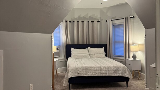 A bedroom with a bed, a nightstand, a lamp, a window with curtains, and a ceiling light. - NH3 - Historic Charm - Cozy 3BR Apartment (DuBois)