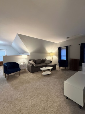 Un salon moderne avec un canapé d’angle, une table basse ronde, un fauteuil bleu, une table d’appoint blanche et une lampe.
