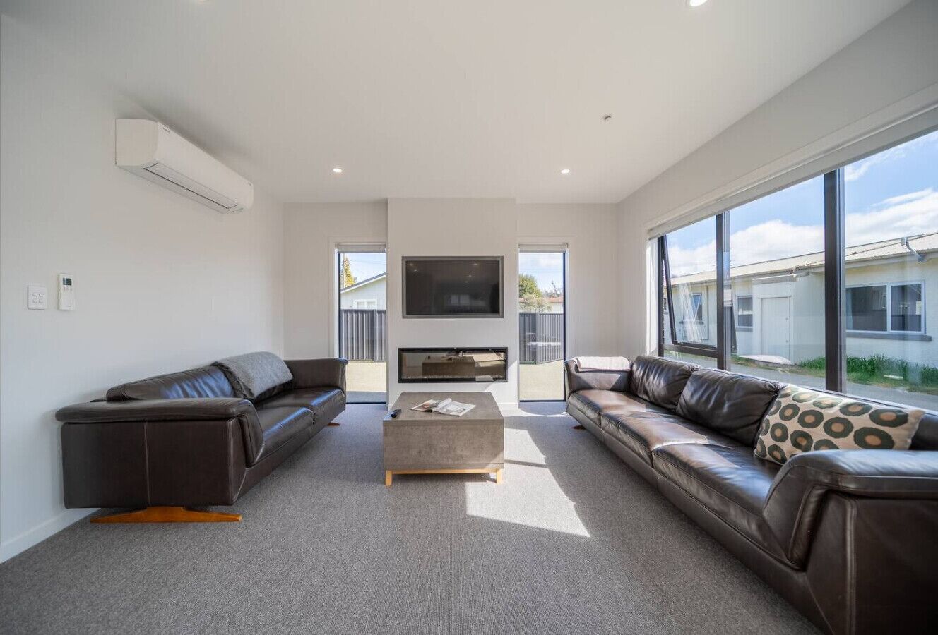A modern living room with a grey carpet, two leather sofas, a coffee table, and a flat-screen TV mounted on the wall.