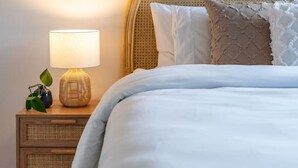 Una cama bien tendida con ropa de cama blanca, un cabecero de mimbre y una lámpara de noche con pantalla blanca.