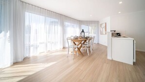 Une pièce spacieuse avec une table et des chaises en bois, un petit coin cuisine et de grandes fenêtres ornées de rideaux légers.