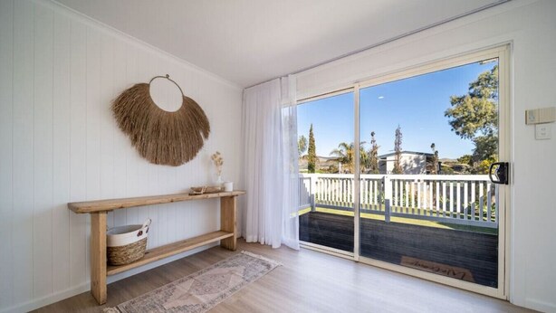 Une pièce avec une console en bois, une décoration murale tissée et une grande porte-fenêtre coulissante donnant sur une terrasse extérieure dotée d’une rambarde blanche.