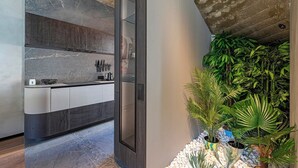 Una cocina moderna con armarios de madera oscura, encimera de mármol y estufa integrada. Hay una pequeña isla con un fregadero y un hervidor. La cocina tiene techo de concreto y un pequeño jardín interior con diversas plantas.