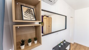 Une chambre moderne avec une étagère en bois sur laquelle sont posés un tableau représentant une feuille encadrée et de petites plantes en pot, un grand miroir qui reflète une armoire et un luminaire suspendu, ainsi qu’une petite table noire agrémentée d’autres plantes.