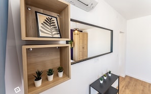 Ein modernes Zimmer mit einem hölzernen Regal, das eine gerahmte Blattform und kleine Topfpflanzen beherbergt, einem großen Spiegel, der eine Garderobe und eine Pendelleuchte reflektiert, sowie einem kleinen schwarzen Tisch mit weiteren Pflanzen.