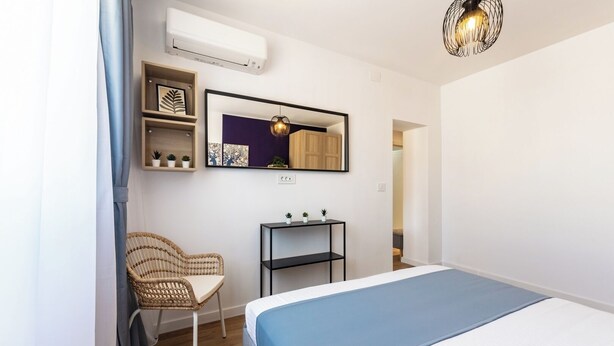 A bedroom with a bed, a chair, a shelf with plants, a mirror, and an air conditioner. - Elegant Apartment in Ivory - AC and Smart TV (Voluntari)