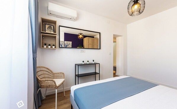 A bedroom with a bed, a chair, a shelf with plants, a mirror, and an air conditioner.