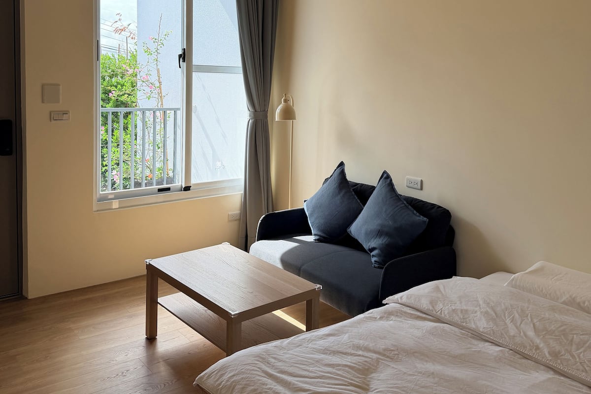 A room with a wooden ceiling, a window with a view of greenery, a sofa with cushions, a bed, and a small table.