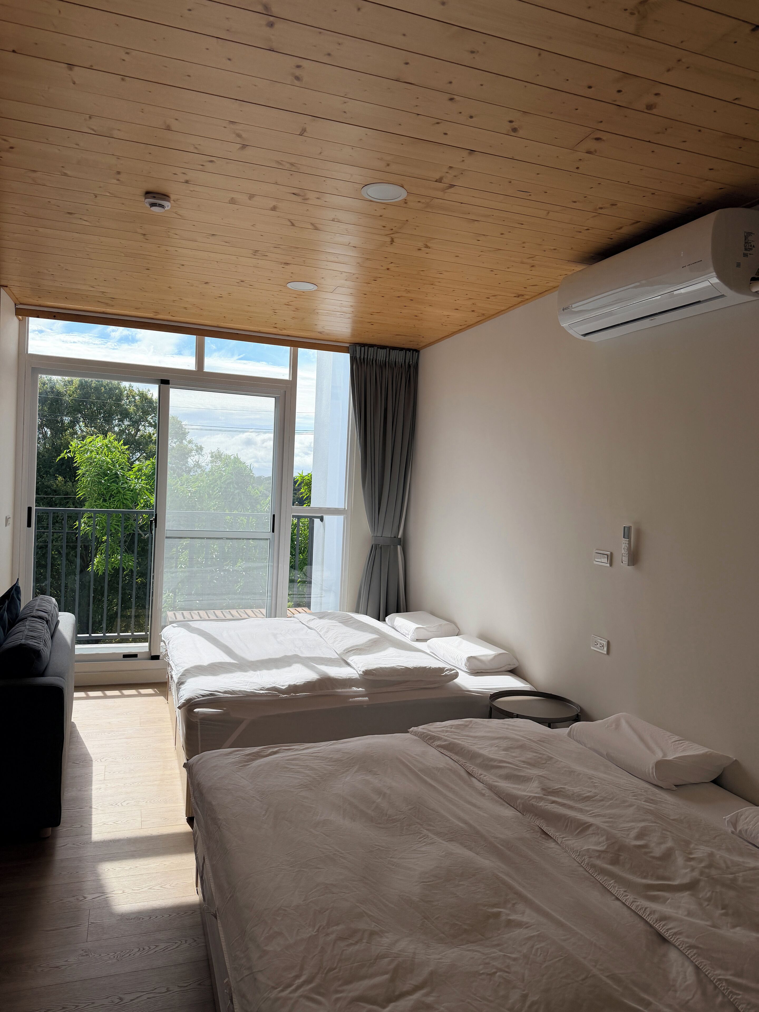 Una habitación de hotel con techo de madera, dos camas, un balcón con vistas y un equipo de aire acondicionado.