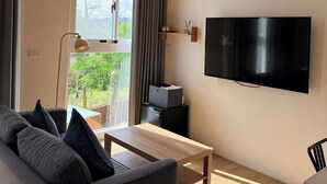 A modern living room with a wooden ceiling, a large flat-screen TV mounted on the wall, a grey sofa, a wooden coffee table, and a view of greenery outside. - KUMOYOKU B&B (Taichung)