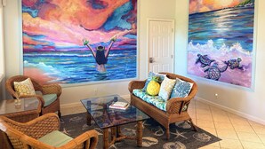 Una habitación con un gran ventanal, muebles de mimbre y dos cuadros grandes que muestran a una persona con los brazos levantados frente a un cielo y mar coloridos.