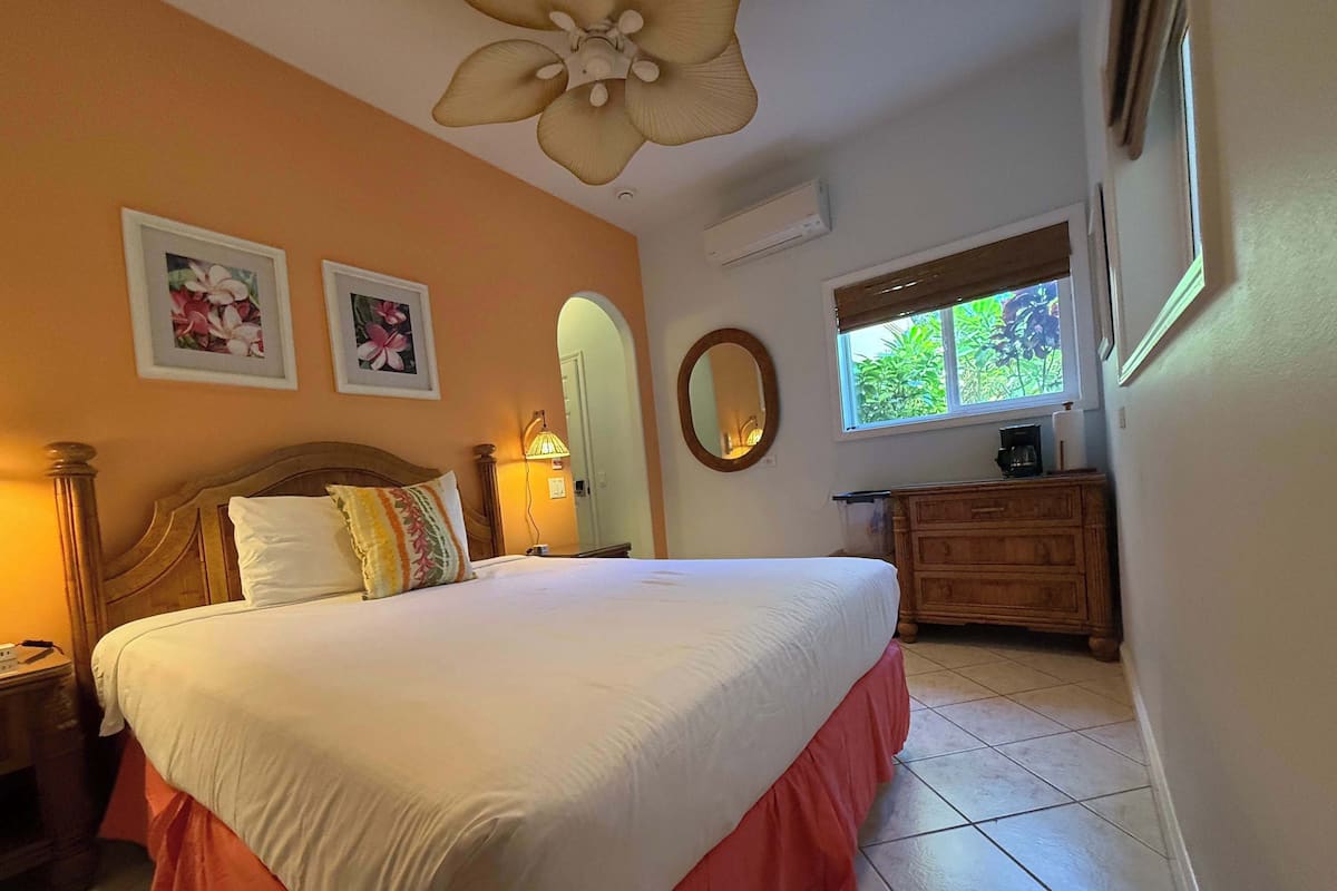A neatly made bed with a wooden headboard, a nightstand with a lamp, a dresser, and a window with a view of greenery.