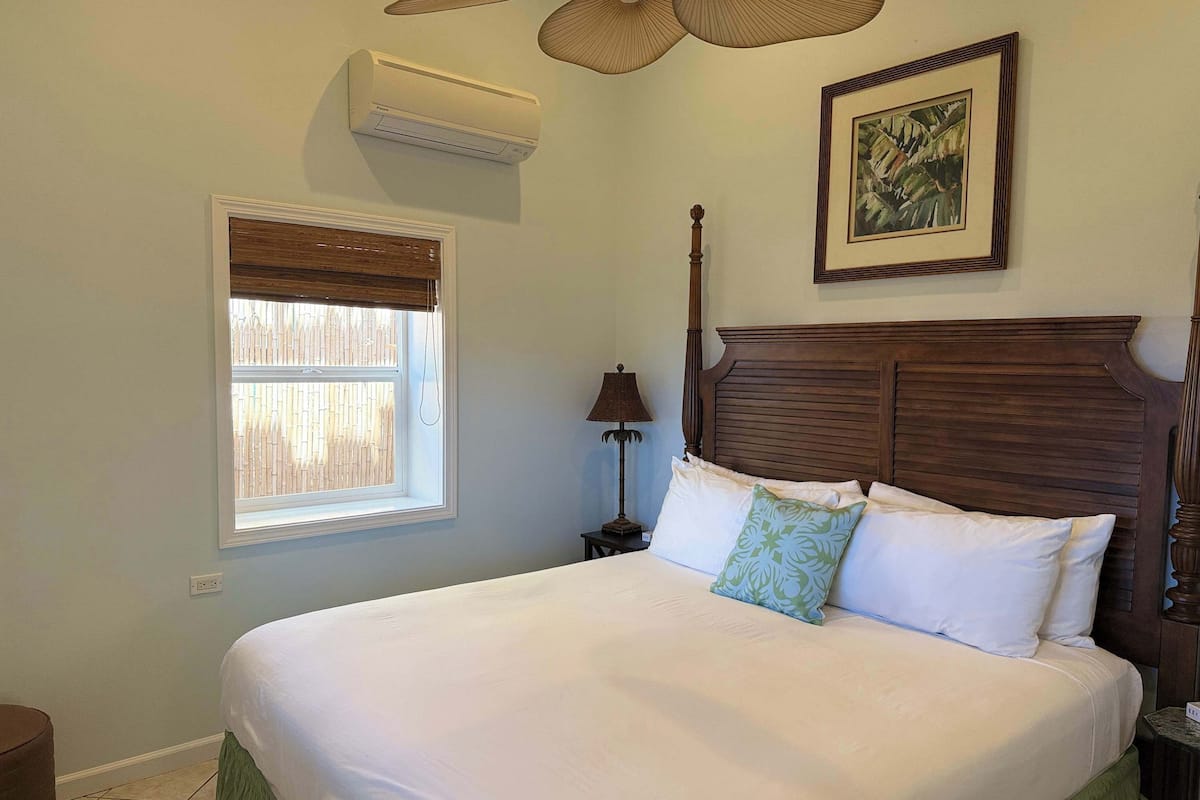A bedroom with a large bed, a window with blinds, an air conditioning unit, a bedside lamp, and a framed picture on the wall.
