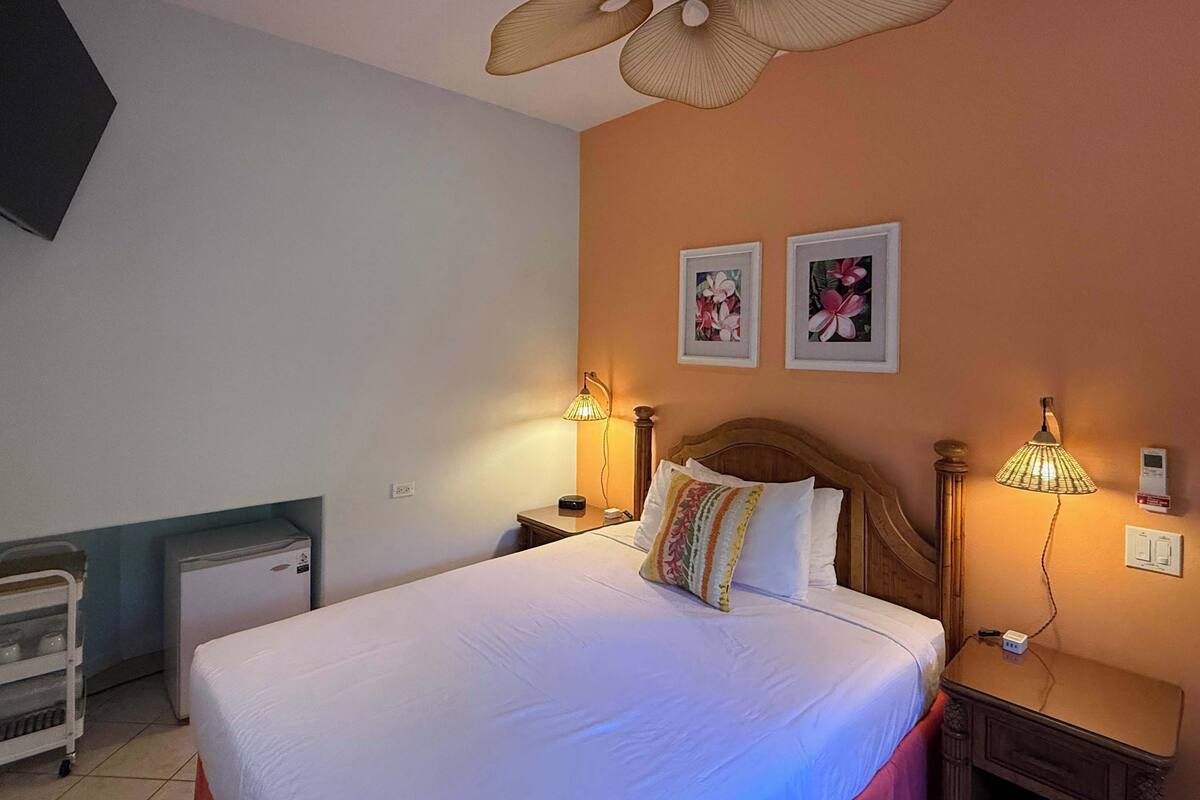 A hotel room with a bed, bedside lamps, a ceiling fan, a small desk, and a refrigerator.