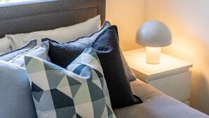 Una cama bien hecha con almohadas de estampado geométrico, una lámpara de noche y una ventana con vistas al verdor.