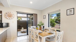 Un coin repas avec une table en bois dressée pour quatre personnes, des chaises blanches, une horloge et une vue sur l’extérieur grâce à une grande fenêtre.