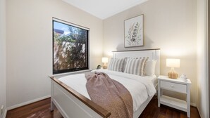 Une chambre avec une grande fenêtre, un lit doté d’une tête de lit blanche, une table de chevet avec une lampe et un tableau encadré accroché au mur.
