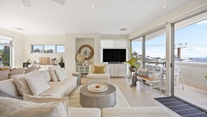 A spacious living room with a large sofa set, a coffee table, and a TV. There is a balcony with a view of the sea.
