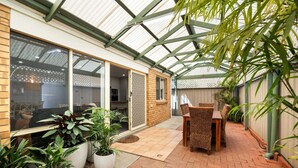A covered patio area with a brick floor, potted plants, wicker furniture, and a view of the adjacent building.