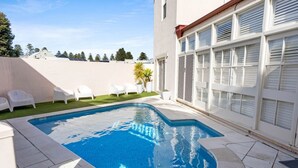 A swimming pool with a tiled deck, white lounge chairs, and a building with large windows and shutters. - Sea Breeze @ Smugglers Inn (Victor Harbor)