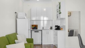 A compact kitchen with white cabinets, a small refrigerator, and a coffee machine. There is a green sofa with a white throw pillow in the living area. - Sea Breeze @ Smugglers Inn (Victor Harbor)