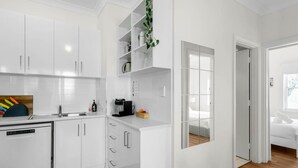 A modern kitchen with white cabinetry, a dishwasher, and a coffee maker. There is a small plant on a shelf and a window with a view of a bedroom. - Sea Breeze @ Smugglers Inn (Victor Harbor)