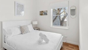 A white bedroom with a bed, a bedside table, a lamp, a window with a tree view, and two framed artworks on the wall. - Sea Breeze @ Smugglers Inn (Victor Harbor)