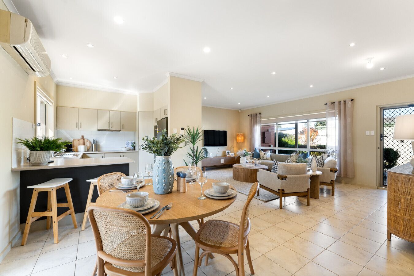 Une cuisine moderne avec des meubles blancs, un coin repas avec une table ronde et des chaises en osier, et un salon avec un canapé et un téléviseur.