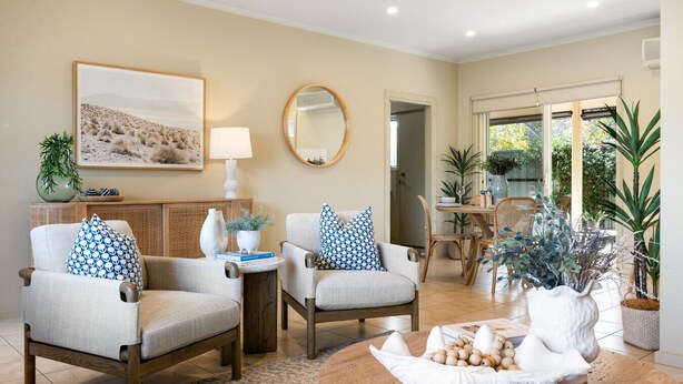 A well-lit living room with two armchairs, a coffee table, a painting, a mirror, and a dining area with a table and chairs.