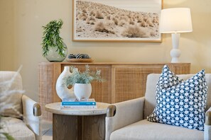 Una sala acogedora con un sofá, una mesita de noche con libros, una lámpara y jarrones decorativos.