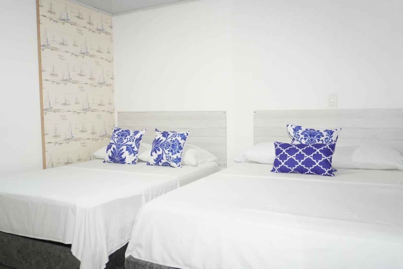 Two single beds with white bedding and blue patterned pillows, a white wall with a sailboat wallpaper, and a wooden headboard.