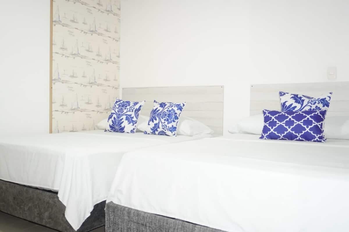 Deux lits simples avec du linge de lit blanc et des oreillers à motifs bleus, un mur blanc recouvert d’un papier peint sur le thème de la marine, et une tête de lit grise.