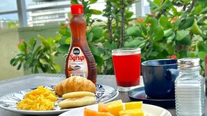 Een ontbijttafel met roerei, croissants en fruit op borden, een fles siroop, een glas rode drank en een blauwe mok.