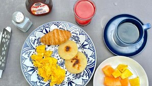 Een bord met roerei, een croissant en twee kleine pannenkoeken, vergezeld van een glas rode sap en een kom blokjes fruit.
