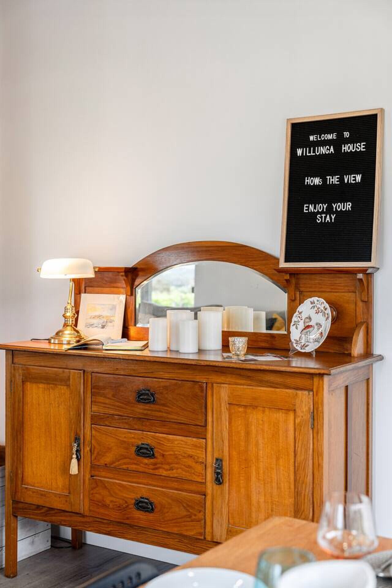 Een houten dressoir met een spiegel, een lamp en een bord waarop staat: WELCOME TO WILLUNGA HOUSE HOW'S THE VIEW ENJOY YOUR STAY.