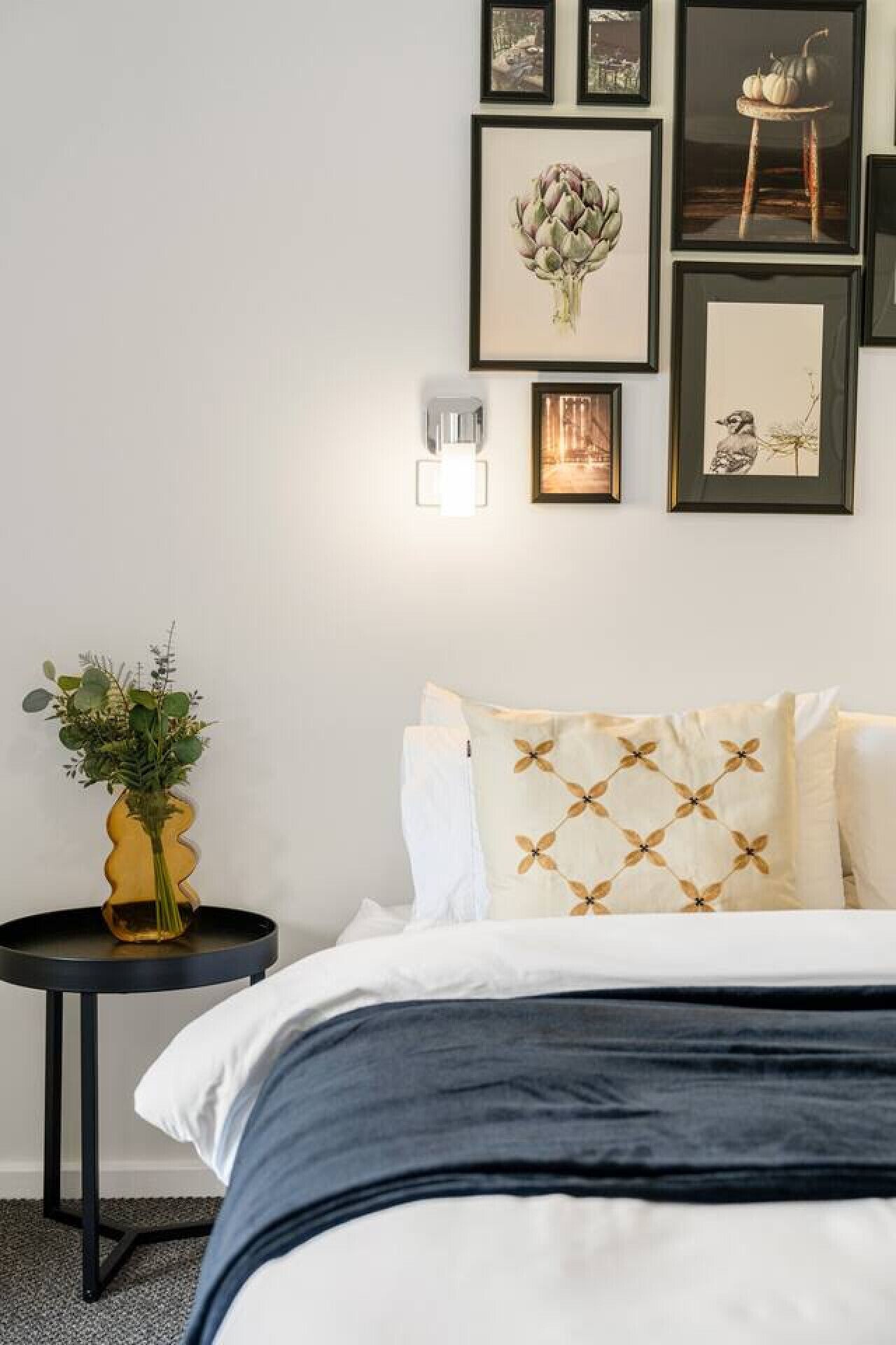 Ein Schlafzimmer mit einem Bett, einem Nachttisch mit Blumenvase und einer Wand mit gerahmten Kunstwerken.
