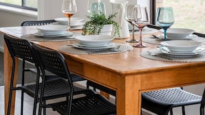 Une table à manger est dressée avec des assiettes, des verres et un pichet, entourée de chaises noires. Une fenêtre et un tableau encadré ornent le mur.