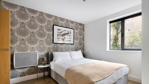 A bedroom with a patterned wallpaper, a bed with white linens, a bedside table with a lamp, and a window with a view of trees. - Blueground | Chalk Farm, terrace, nr Primrose Hill (London)