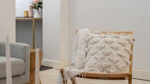 Une pièce au décor minimaliste, avec un canapé gris, une chaise en bois recouverte d’un plaid blanc et une table ronde agrémentée de livres et de bougies.