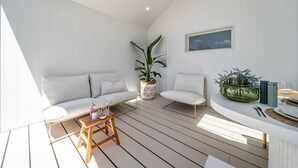 Un intérieur lumineux et blanc, avec un plancher en bois, un canapé, un fauteuil, une petite table sur laquelle se trouvent une bouteille et des verres, et une plante en pot.