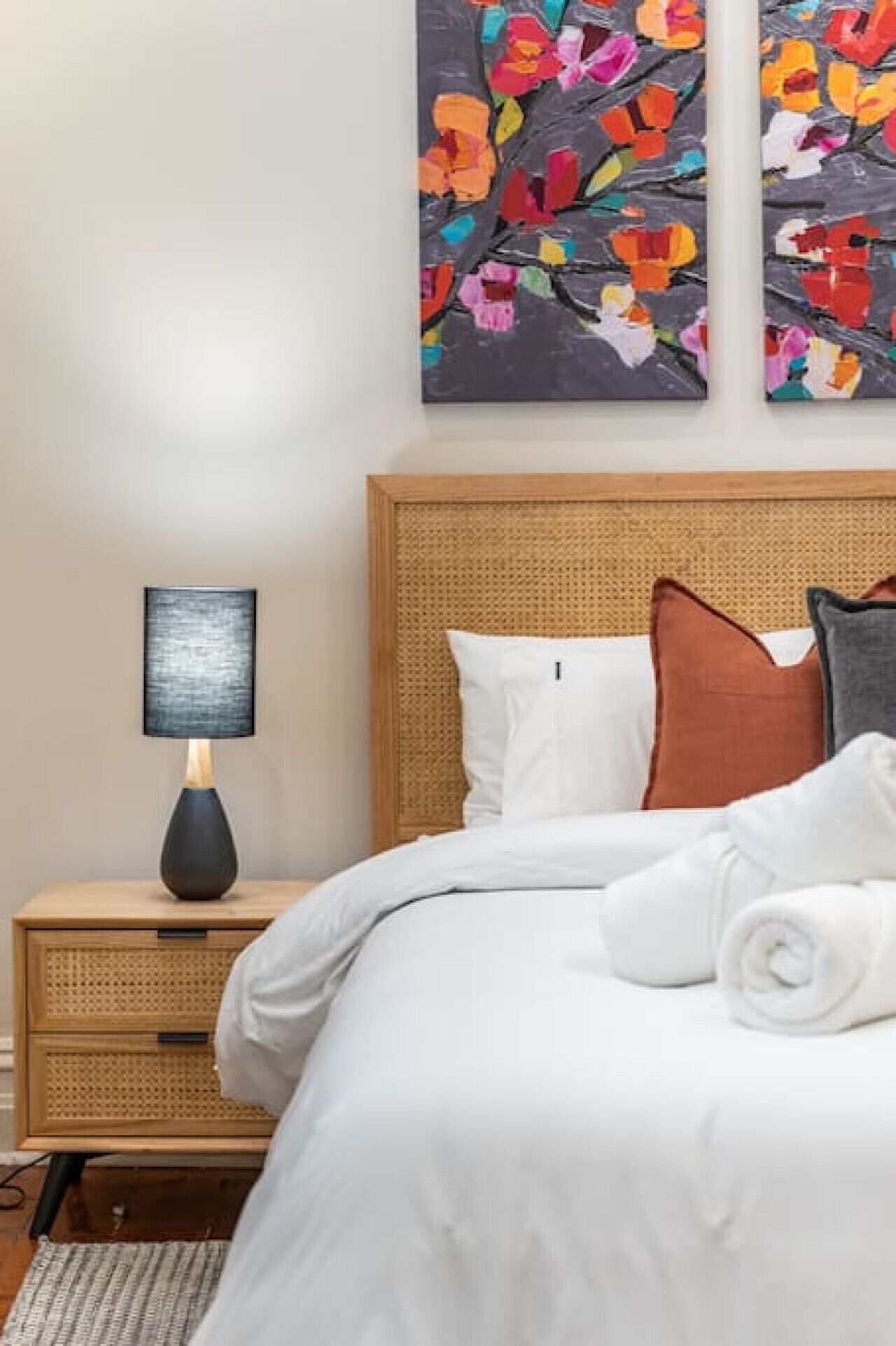 A neatly made bed with white bedding, a wooden nightstand with a lamp, and two colorful abstract paintings on the wall.