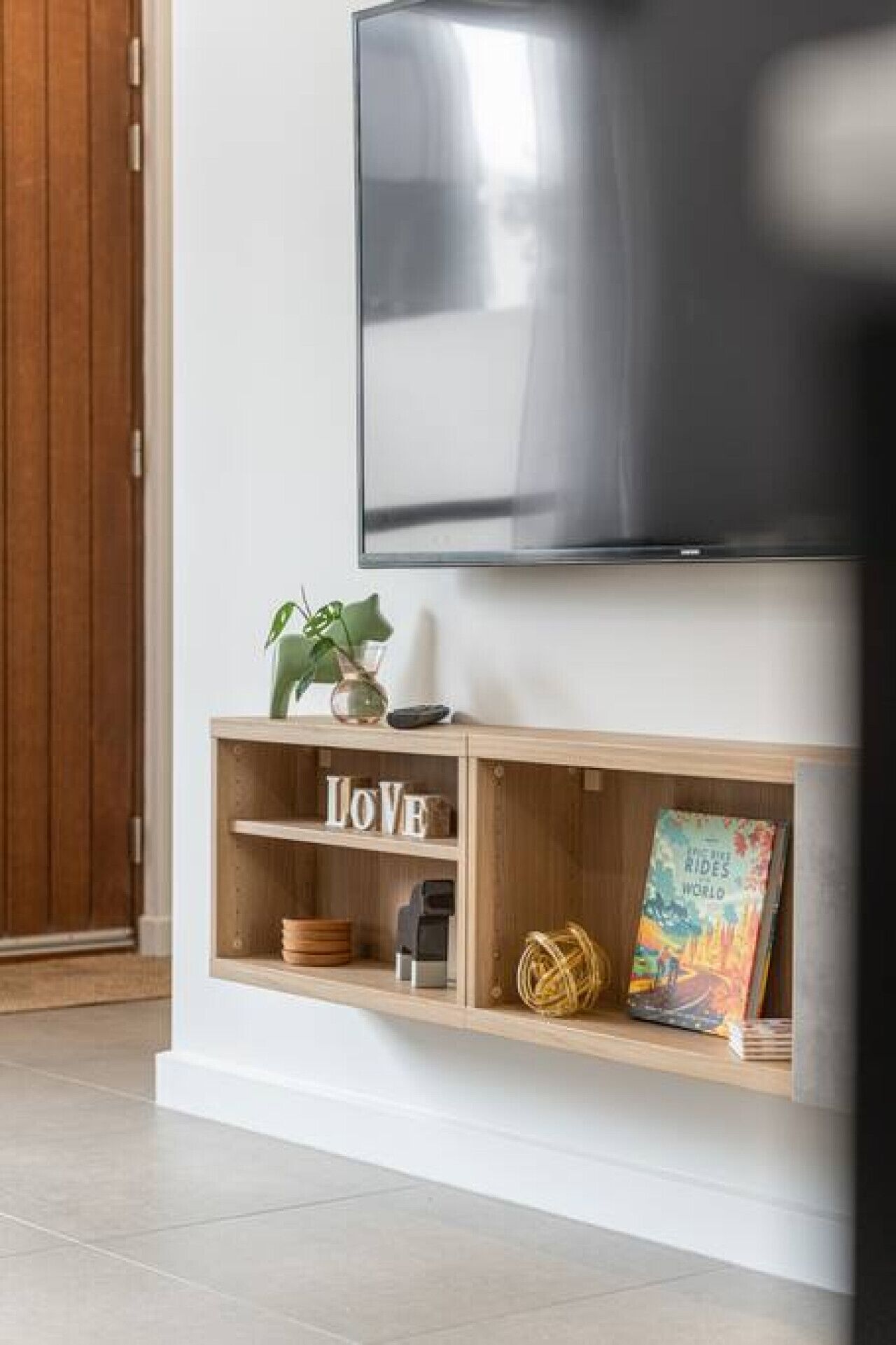 A modern living room with a flat-screen TV mounted on the wall, a wooden console table with decorative items, and a large wooden door.